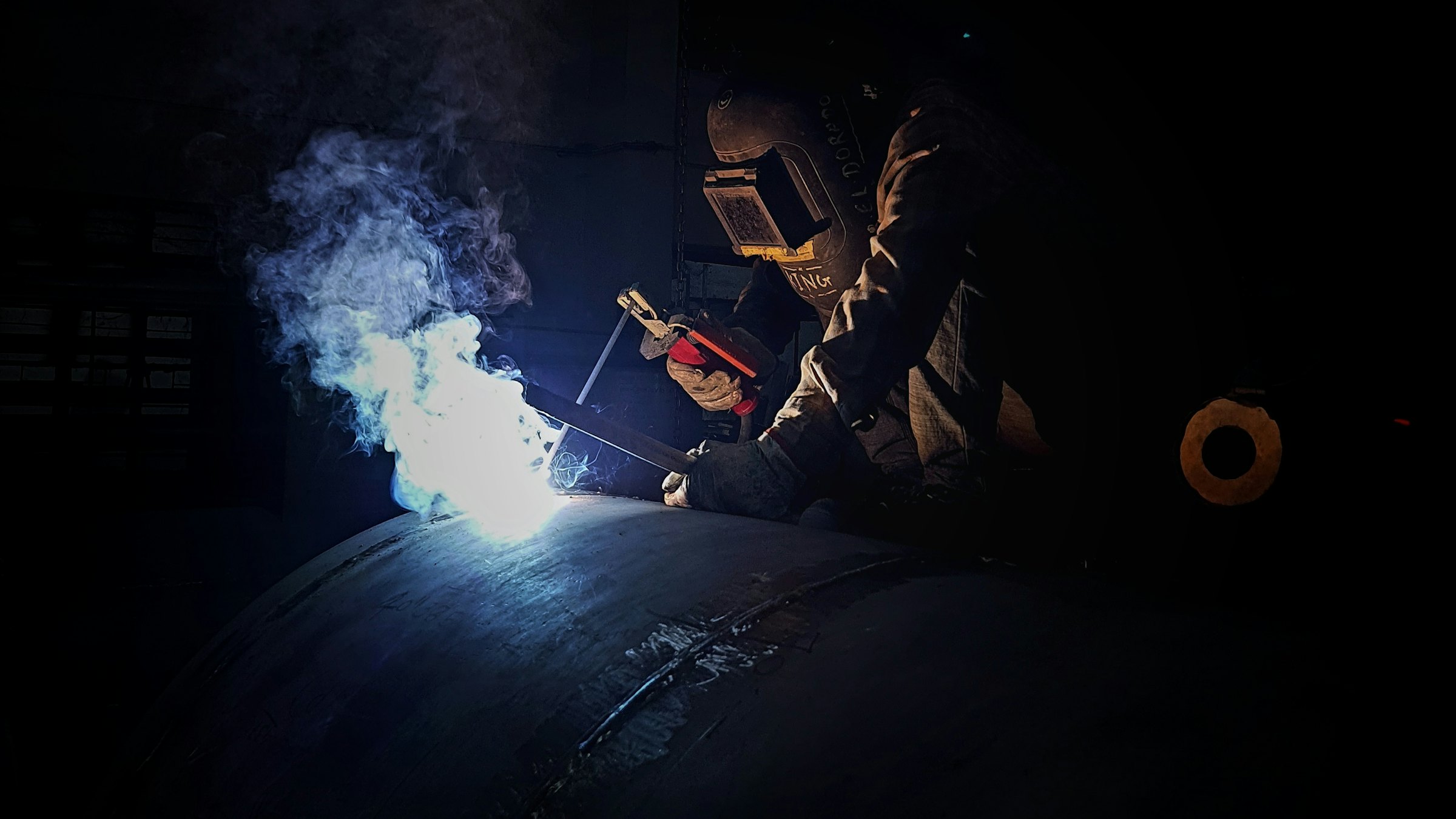 A welder joins a steel pipe in the dark, the welding arc lighting up smoke and the pipe surface
